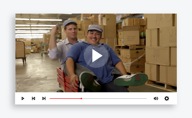Paused video image of a two men riding in a wagon in front of stacks of boxes.