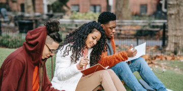 Three college students sitting outside on a college campus talking and taking ntoes.