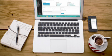 Laptop open on wood table with coffee, cellphone, and open notepad.