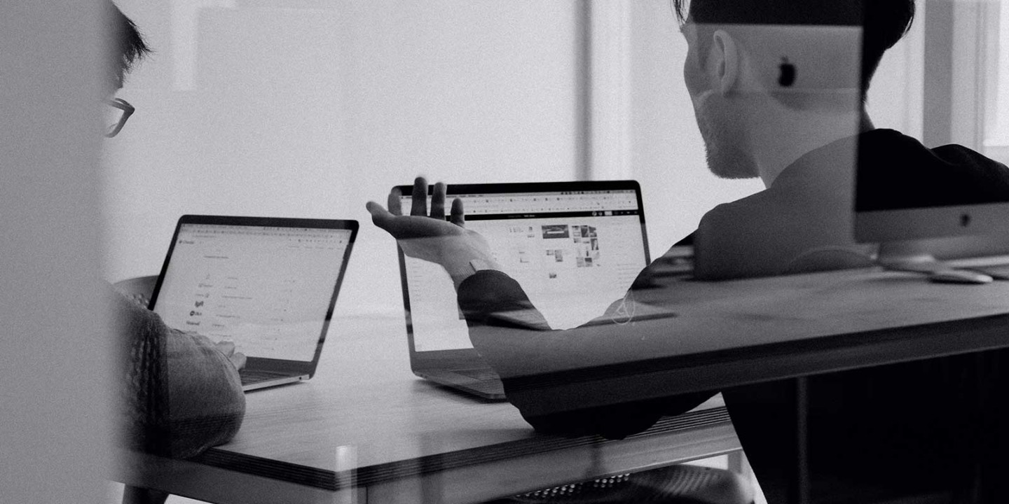 Black and white photo of two people talking in front of laptops.
