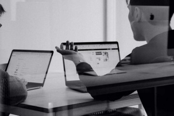 Black and white photo of two people talking in front of laptops.