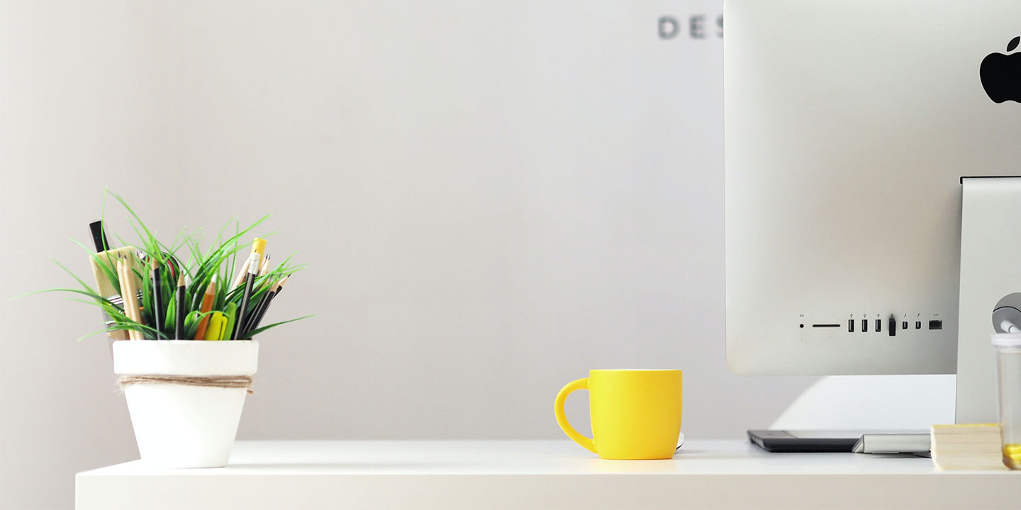 Apple computer on desk with yellow mug and small green plant.