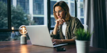 A woman learning about customer journey mapping with shoulder-length hair wearing glasses sits at a desk looking at a laptop with a large window behind her.