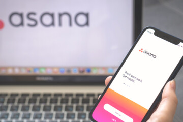 A close up of a person holding a phone with Asana on the screen in front of a laptop with Asana on the screen.