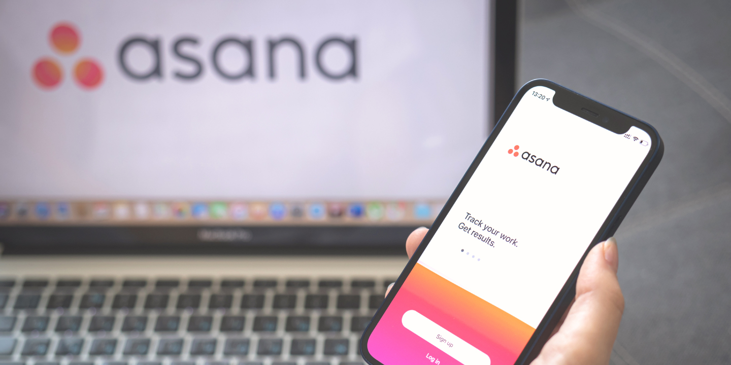 A close up of a person holding a phone with Asana on the screen in front of a laptop with Asana on the screen.