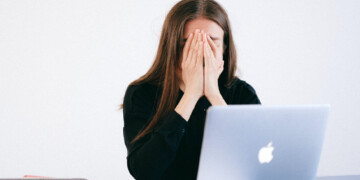 A woman holds her head her hands while sitting in front of an Apple laptop.