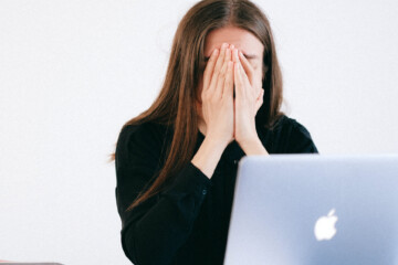 A woman holds her head her hands while sitting in front of an Apple laptop.