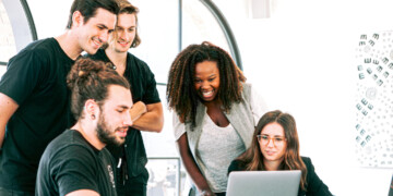 A group of five people gather around a lap top in front of a window.