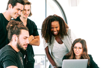 A group of five people gather around a lap top in front of a window.