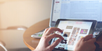 Close up of a person scrolling on an iPad in front of a laptop.