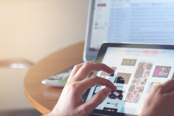 Close up of a person scrolling on an iPad in front of a laptop.