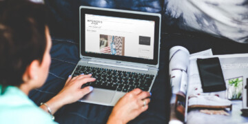 A person types on their laptop with an open magazine and cell phone to their right.