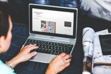 A person types on their laptop with an open magazine and cell phone to their right.