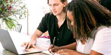 One person looks at a laptop while another person writes notes in a notebook.