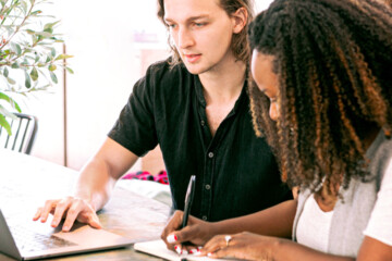 One person looks at a laptop while another person writes notes in a notebook.