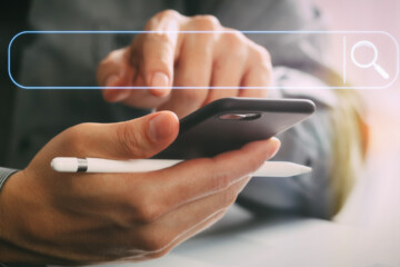 A person poises to touch the screen of a cell phone while holding a white pen.