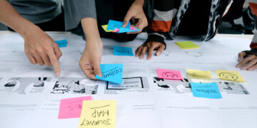 A group of coworkers stands at a table as they they develop a customer journey map while sharing notes to track their progress.