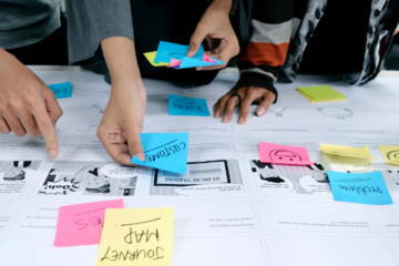 A group of coworkers stands at a table as they they develop a customer journey map while sharing notes to track their progress.