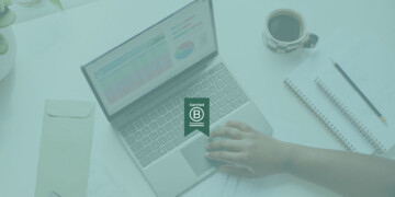 A certified B Corp badge logo overlaid on top of a background image of someone using their laptop.