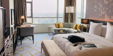 Hotel room with a picture window and blinds, couch with yellow cushions, and queen bed with beige covers and pillows.