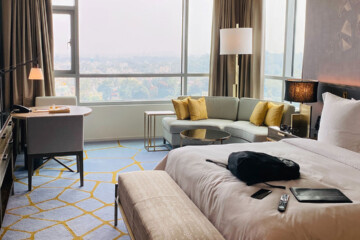 Hotel room with a picture window and blinds, couch with yellow cushions, and queen bed with beige covers and pillows.