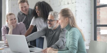 A group of coworkers sit at a table smiling while looking at a laptop computer.