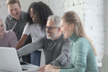 A group of coworkers sit at a table smiling while looking at a laptop computer.