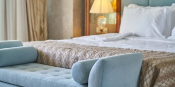 A luxe hotel room features a king bed with crisp white sheets, fluffy pillows, and patterned gold colored comforter as well as a light blue upholstered end-of-bed bench.