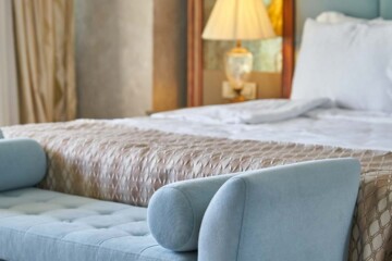 A luxe hotel room features a king bed with crisp white sheets, fluffy pillows, and patterned gold colored comforter as well as a light blue upholstered end-of-bed bench.
