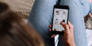 A young woman with brown hair and wearing blue jeans sits on her couch while scrolling through ads on her smartphone.