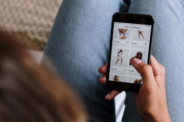 A young woman with brown hair and wearing blue jeans sits on her couch while scrolling through ads on her smartphone.