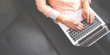 A woman with red manicured nail has her hands on her laptop while sitting on a couch.