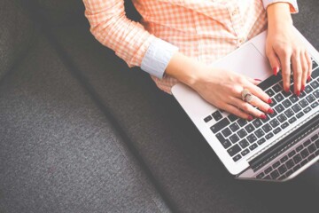 A woman with red manicured nail has her hands on her laptop while sitting on a couch.