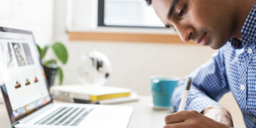 A man in a blue and white checkered shirt sits at a desk in front of his laptop while writing on a notepad.