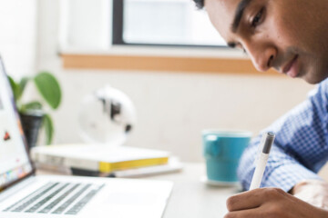 A man in a blue and white checkered shirt sits at a desk in front of his laptop while writing on a notepad.