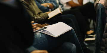 A women sitting at a conference while taking notes with a notebook, pen and her phone.