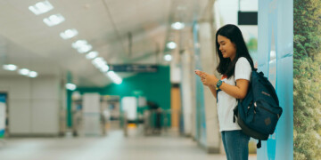 An Asian college student with a blue backpack stands in a school hallway while looking at her cell phone.