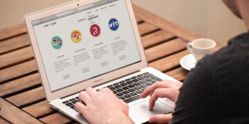 A man sits at a wood table with a cup of espresso and types on his laptop.