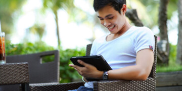A young man in a white t-shirt sits at a patio table drinking iced tea while scrolling through an iPad.