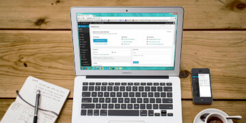 Laptop on a wood desk with pad of paper, pen, smartphone, and cup of coffee.