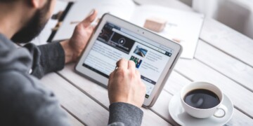 Person using a tablet to browse the web, with a cup of coffee nearby on a wooden table.