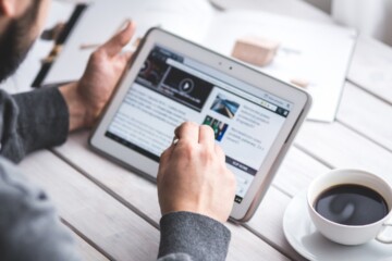 Person using a tablet to browse the web, with a cup of coffee nearby on a wooden table.