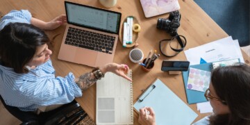 Two female coworkers sitting at a wood desk with laptop computer, calendars, camera, paper, and pens.