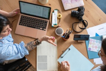 Two female coworkers sitting at a wood desk with laptop computer, calendars, camera, paper, and pens.