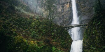 Multnomah Falls cascading down moss-covered cliffs with the iconic Benson Bridge spanning across the waterfall, surrounded by lush green forest near Portland, Oregon