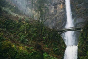 Multnomah Falls cascading down moss-covered cliffs with the iconic Benson Bridge spanning across the waterfall, surrounded by lush green forest near Portland, Oregon