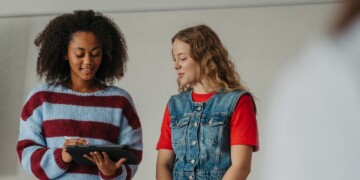 Two young women standing together and looking at a tablet, one wearing a striped sweater and the other a denim vest over a red shirt.