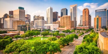 Aerial view of the downtown Houston, Texas skyline with lush green trees and park space in the foreground on a sunny day.